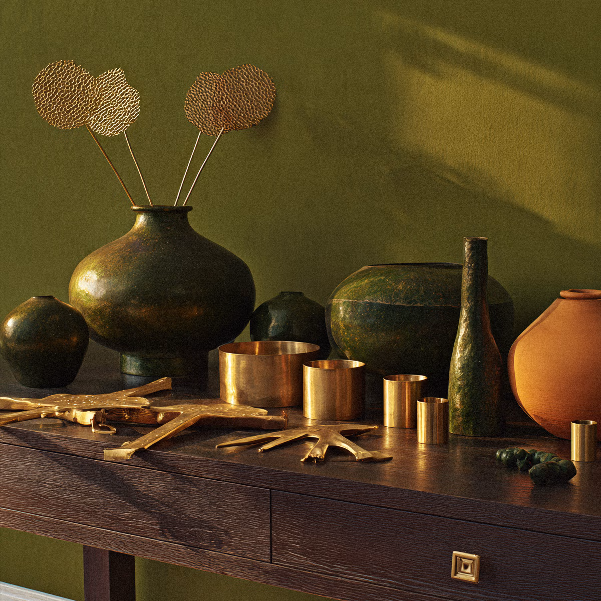 Collection d'objets décoratifs sur console en bois wengé avec vases en céramique aux patines vert-de-gris oxydé, récipients cylindriques en laiton doré brossé, jarre terracotta et sculptures décoratives dorées, sur fond de mur vert olive profond éclairé par lumière rasante révélant textures et reflets