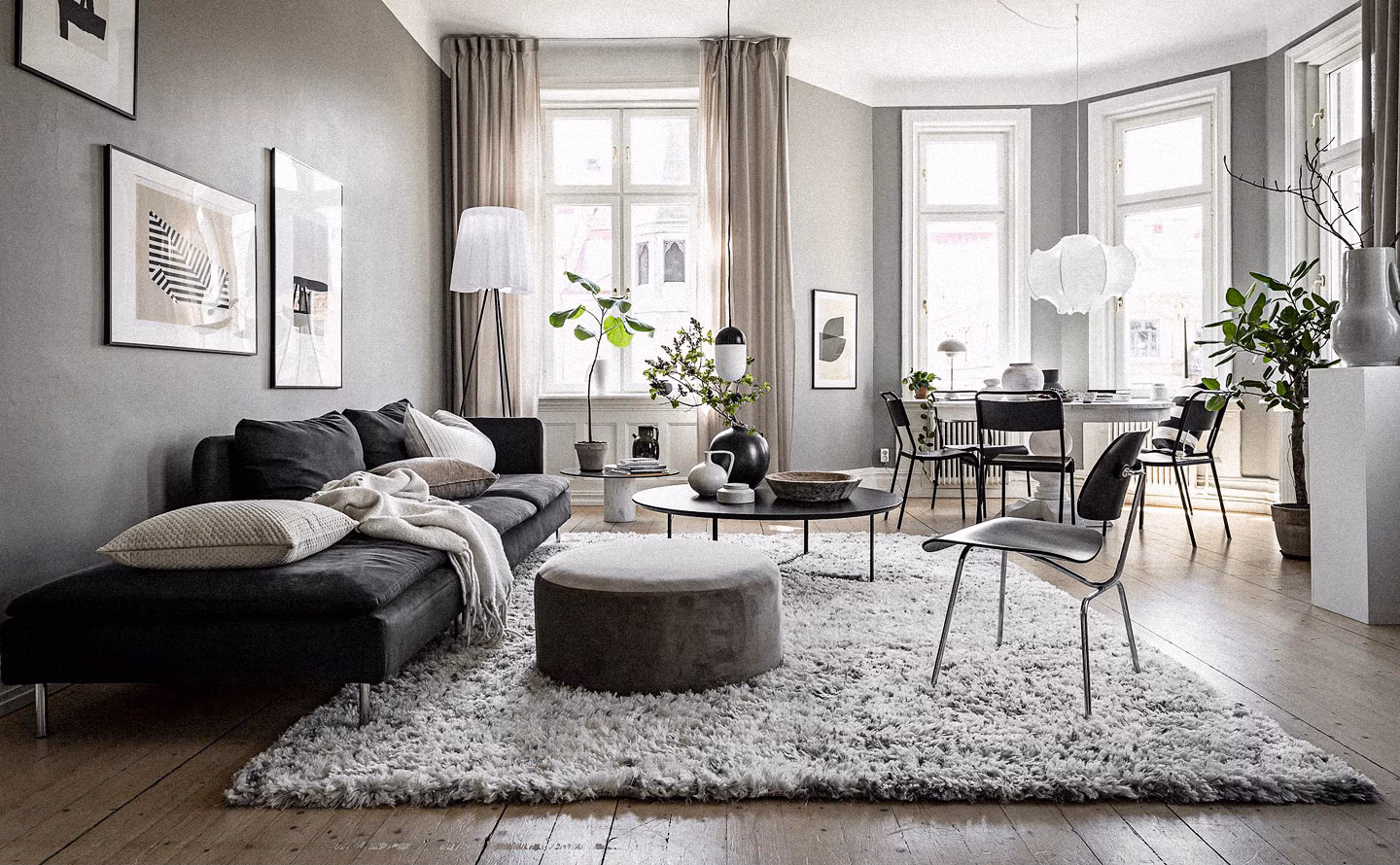 alon-salle à manger ouvert style scandinave contemporain avec canapé d'angle en velours gris anthracite, pouf rond taupe, tapis shaggy gris clair texturé, table basse ronde noire, murs gris perle, parquet chêne blanchi, grandes fenêtres aux rideaux lin et plantes vertes apportant fraîcheur à l'ensemble