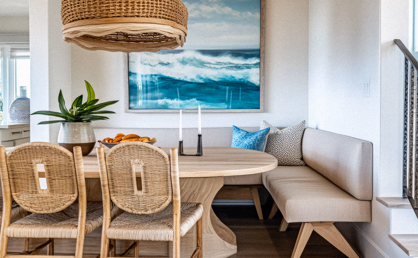 Coin repas côtier avec banquette beige en L, table ronde en bois clair, chaises en rotin naturel, suspension tressée et grand tableau marine illustrant l'intimité d'un espace réduit mais convivial Coin repas côtier avec banquette beige en L, table ronde en bois clair, chaises en rotin naturel, suspension tressée et grand tableau marine illustrant l'intimité d'un espace réduit mais convivial