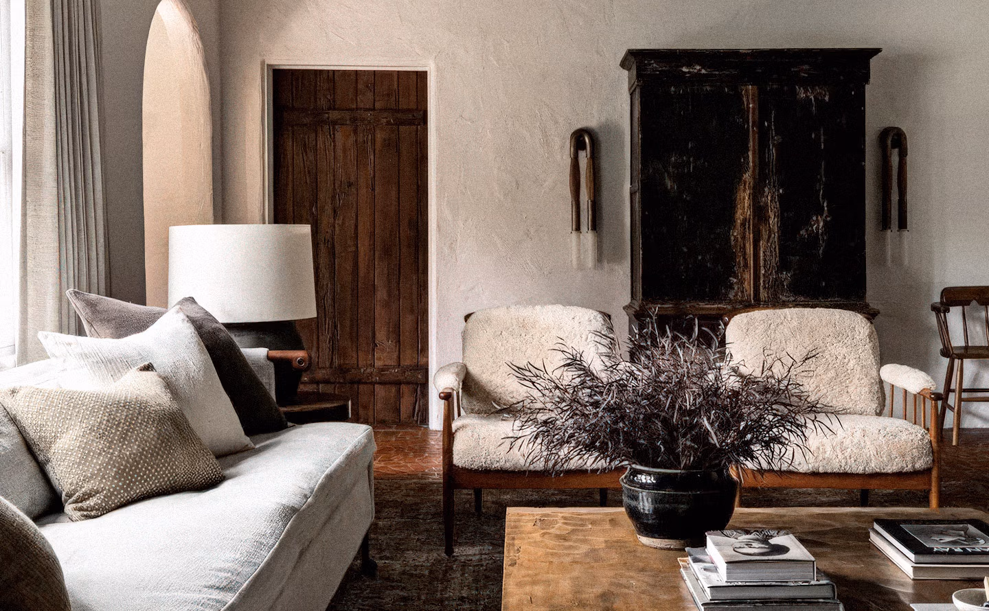 Salon aux murs taupes texturés mêlant mobilier ancien (armoire laquée noire patinée, banquette bois vintage, porte brune ancienne) et contemporain (canapé bouclette beige), avec table basse bois brut, branches séchées en pot noir et textiles naturels créant une atmosphère wabi-sabi raffinée où dialoguent les époques. Salon aux murs taupes texturés mêlant mobilier ancien (armoire laquée noire patinée, banquette bois vintage, porte brune ancienne) et contemporain (canapé bouclette beige), avec table basse bois brut, branches séchées en pot noir et textiles naturels créant une atmosphère wabi-sabi raffinée où dialoguent les époques.