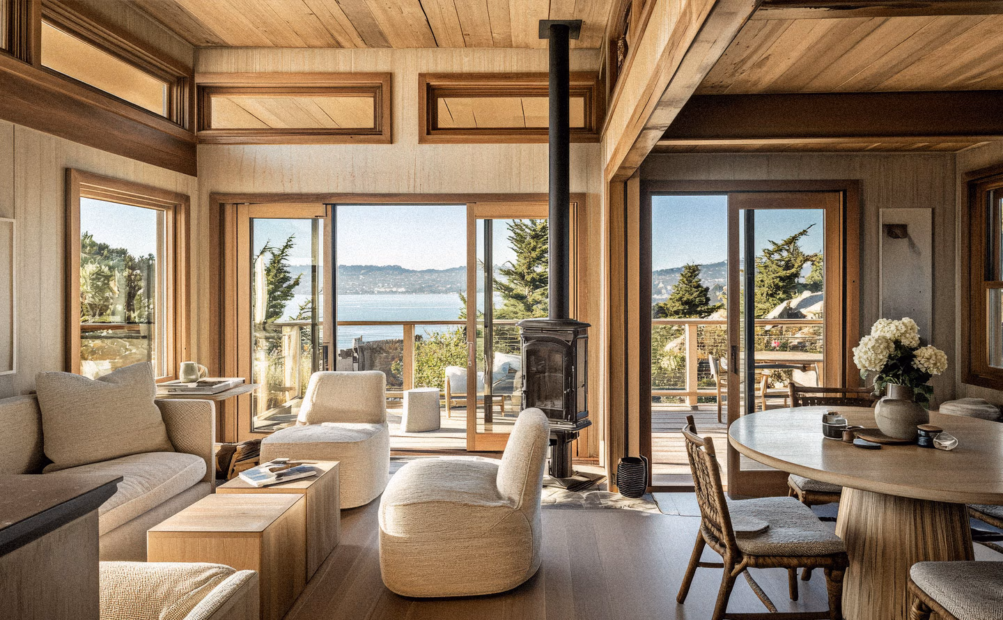 Salon contemporain chaleureux avec plafond et structure en bois clair, grandes baies vitrées panoramiques encadrant vue spectaculaire sur baie maritime, poêle à bois noir central, mobilier beige lin illustrant la transformation réussie d'une maison délabrée en espace de réception valorisant les atouts du site Salon contemporain chaleureux avec plafond et structure en bois clair, grandes baies vitrées panoramiques encadrant vue spectaculaire sur baie maritime, poêle à bois noir central, mobilier beige lin illustrant la transformation réussie d'une maison délabrée en espace de réception valorisant les atouts du site