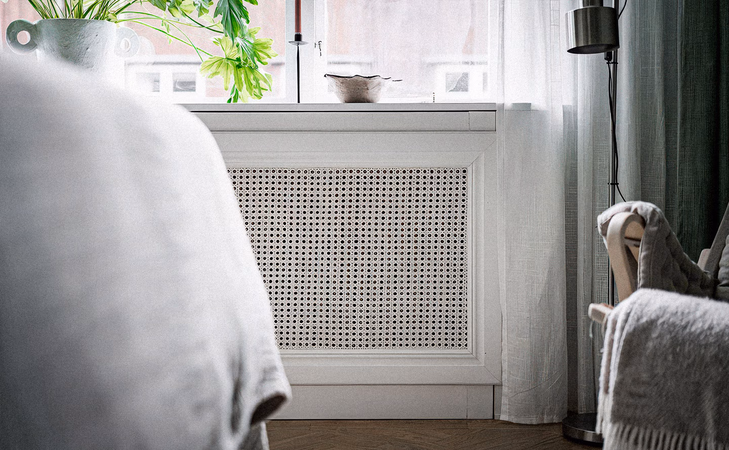 Radiateur blanc dissimulé sous élégant panneau de cannage en rotin naturel tressé, surmonté d'une console blanche étroite présentant vase céramique blanc et plantes vertes luxuriantes, rideaux voilage blanc diffusant lumière douce, lampadaire noir minimaliste et fauteuil velours visible en arrière-plan créant un espace où la contrainte technique devient atout décoratif. Radiateur blanc dissimulé sous élégant panneau de cannage en rotin naturel tressé, surmonté d'une console blanche étroite présentant vase céramique blanc et plantes vertes luxuriantes, rideaux voilage blanc diffusant lumière douce, lampadaire noir minimaliste et fauteuil velours visible en arrière-plan créant un espace où la contrainte technique devient atout décoratif.