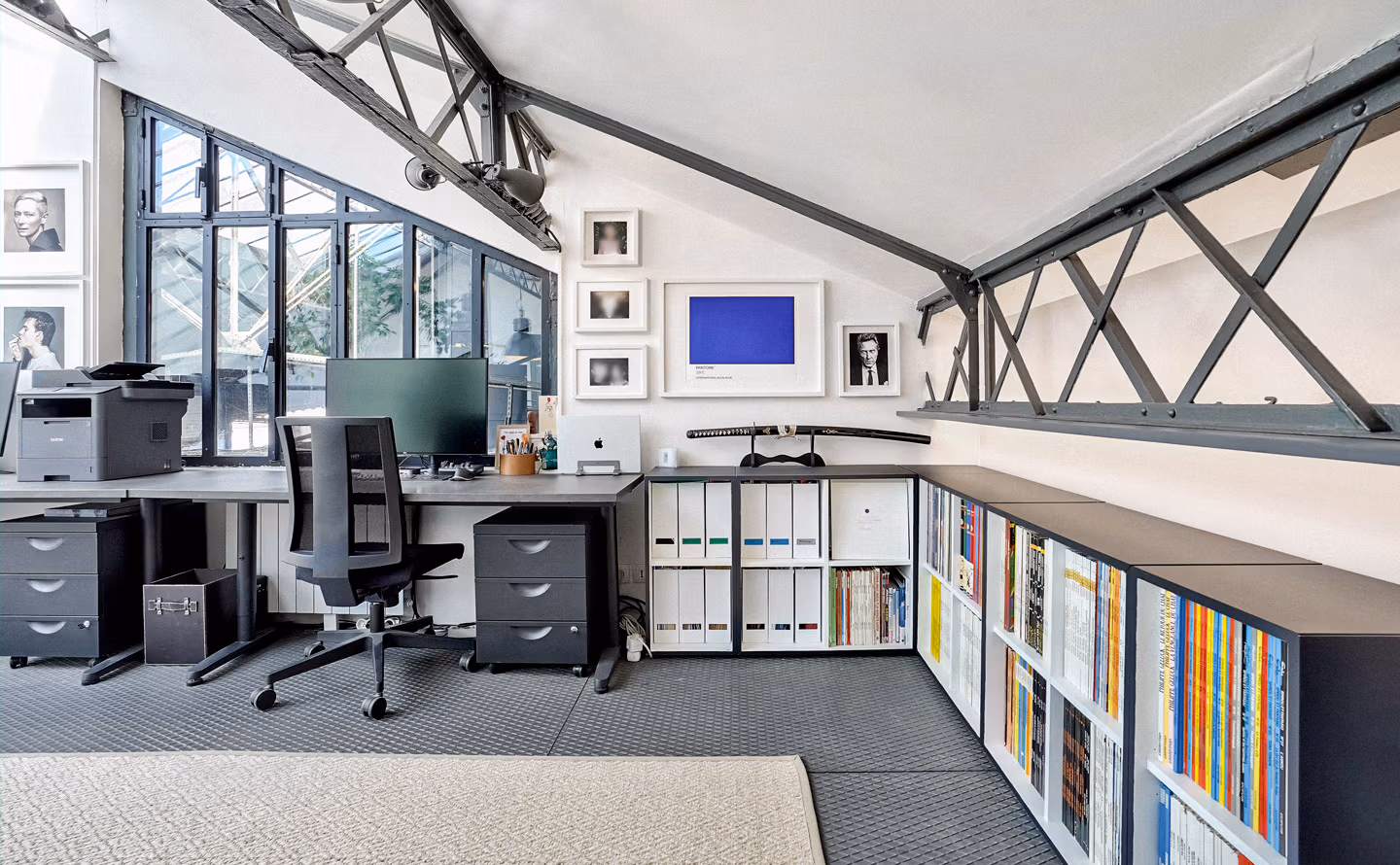 Bureau sous toiture aménagé avec structure métallique apparente en croix, grande verrière d'atelier donnant sur verdure, double poste de travail gris anthracite avec chaises ergonomiques noires, bibliothèque basse blanche présentant livres colorés et classeurs blancs, galerie de portraits noir et blanc avec nuancier Pantone bleu encadré, créant un sanctuaire créatif lumineux et organisé. Bureau sous toiture aménagé avec structure métallique apparente en croix, grande verrière d'atelier donnant sur verdure, double poste de travail gris anthracite avec chaises ergonomiques noires, bibliothèque basse blanche présentant livres colorés et classeurs blancs, galerie de portraits noir et blanc avec nuancier Pantone bleu encadré, créant un sanctuaire créatif lumineux et organisé.