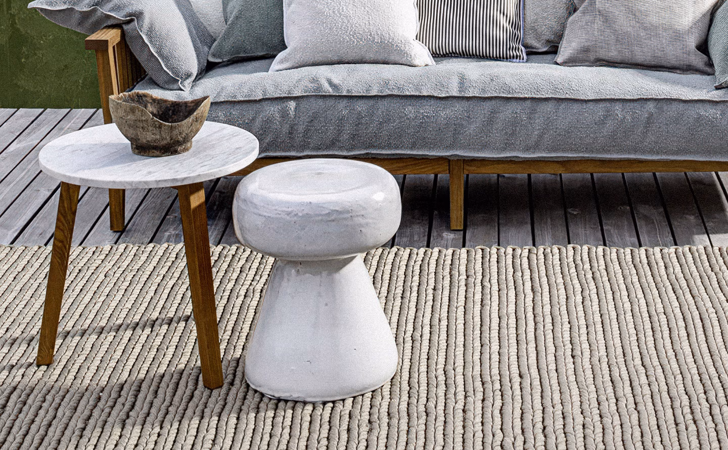 Terrasse extérieure zen avec canapé gris habillé de coussins variés, table basse ronde en marbre blanc veiné sur pieds bois avec bol rustique, tabouret céramique blanc style champignon servant de table d'appoint, épais tapis tressé beige à larges côtes, sol en bois grisé et mur végétal vert illustrant la polyvalence des assises céramiques d'extérieur multifonctions. Terrasse extérieure zen avec canapé gris habillé de coussins variés, table basse ronde en marbre blanc veiné sur pieds bois avec bol rustique, tabouret céramique blanc style champignon servant de table d'appoint, épais tapis tressé beige à larges côtes, sol en bois grisé et mur végétal vert illustrant la polyvalence des assises céramiques d'extérieur multifonctions.