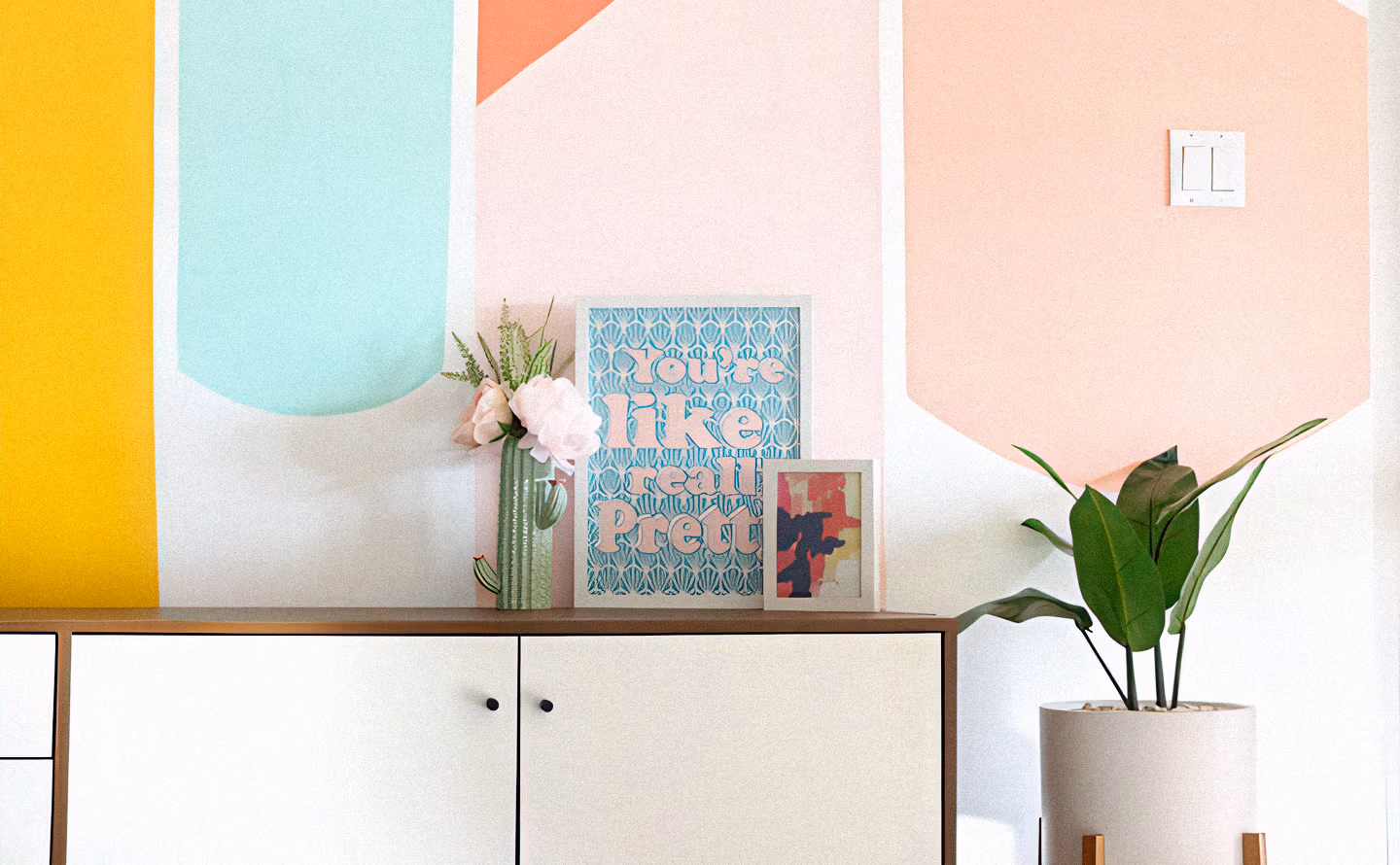 Mur personnalisé avec formes géométriques peintes aux couleurs pastel vives (jaune soleil, bleu ciel, corail, rose pêche), console blanche surmontée de livres colorés empilés, vase rose avec pivoines blanches et cactus, cadre photo, plante oiseau de paradis en pot beige cuivré, illustrant la personnalisation murale réversible par la peinture qui transforme l'espace sans modification structurelle définitive. Mur personnalisé avec formes géométriques peintes aux couleurs pastel vives (jaune soleil, bleu ciel, corail, rose pêche), console blanche surmontée de livres colorés empilés, vase rose avec pivoines blanches et cactus, cadre photo, plante oiseau de paradis en pot beige cuivré, illustrant la personnalisation murale réversible par la peinture qui transforme l'espace sans modification structurelle définitive.
