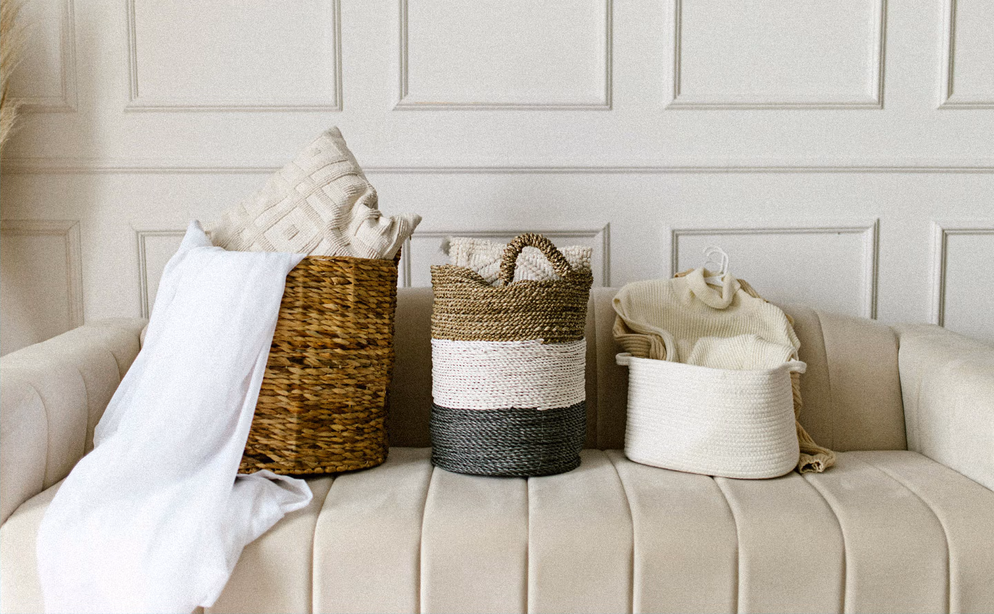 Trois paniers en osier naturel et bicolore sur canapé capitonné beige, textiles lin blanc et beige, mur boiseries classiques blanches, rangement décoratif Trois paniers en osier naturel et bicolore sur canapé capitonné beige, textiles lin blanc et beige, mur boiseries classiques blanches, rangement décoratif