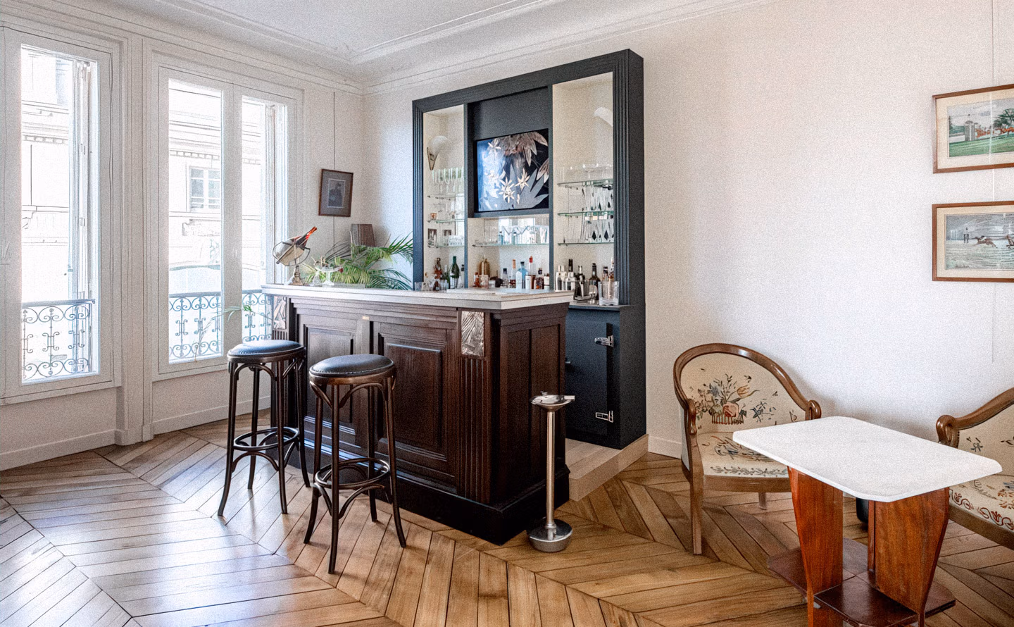 Bar intégré dans un salon haussmannien avec structure noire encadrant miroir et étagères vitrées exposant une collection de spiritueux, comptoir en bois foncé et marbre, tabourets bistrot noirs en cuir, parquet point de Hongrie et moulures d'origine illustrant qu'un véritable bar constitue le point névralgique des réceptions. Bar intégré dans un salon haussmannien avec structure noire encadrant miroir et étagères vitrées exposant une collection de spiritueux, comptoir en bois foncé et marbre, tabourets bistrot noirs en cuir, parquet point de Hongrie et moulures d'origine illustrant qu'un véritable bar constitue le point névralgique des réceptions.