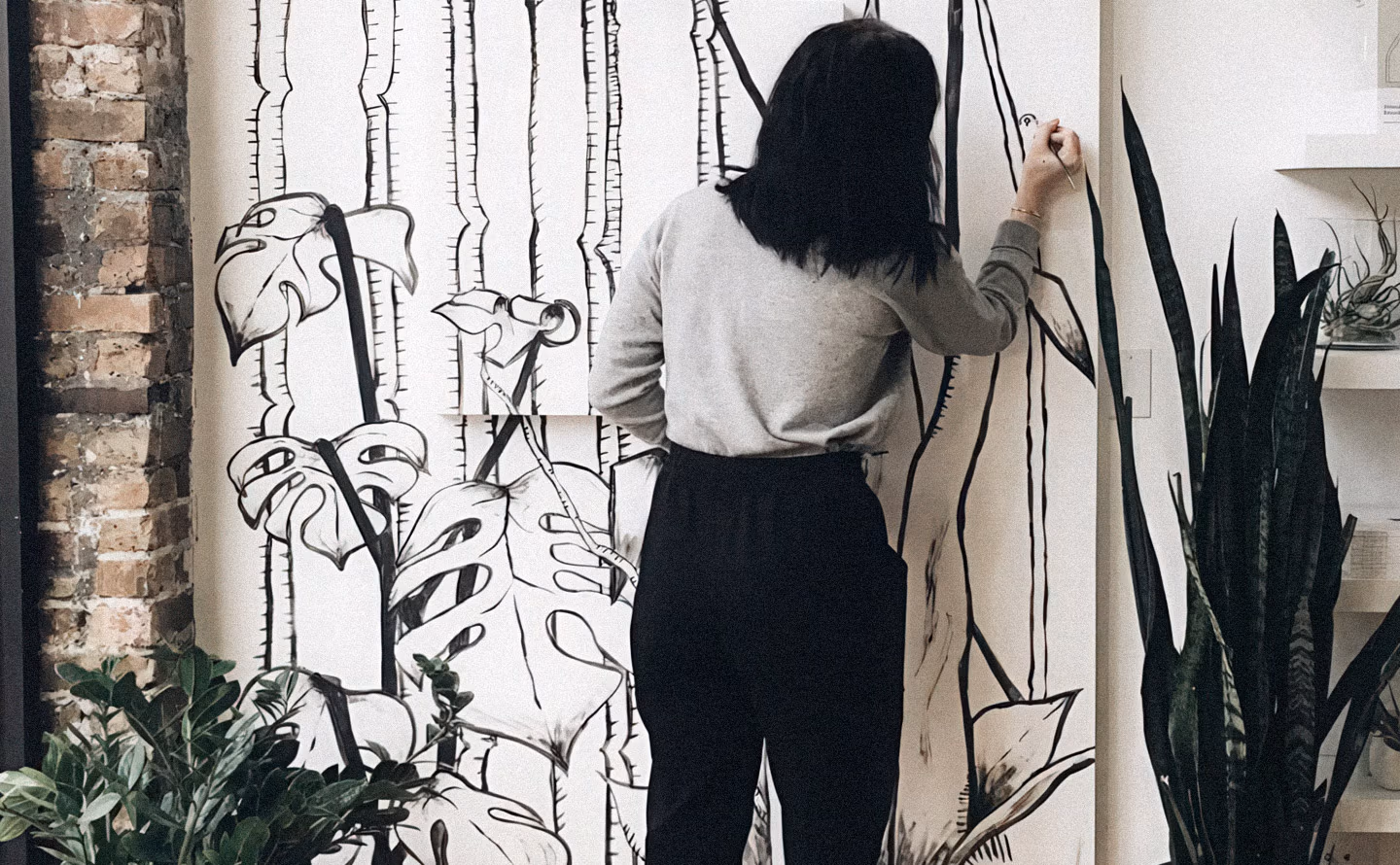 Personne peignant une fresque murale graphique en noir et blanc directement sur un mur avec motifs organiques et lignes à main levée dans un loft aux briques apparentes, entouré de plantes sansevieria, illustrant la transformation décorative DIY accessible avec simplement peinture et audace créative. Personne peignant une fresque murale graphique en noir et blanc directement sur un mur avec motifs organiques et lignes à main levée dans un loft aux briques apparentes, entouré de plantes sansevieria, illustrant la transformation décorative DIY accessible avec simplement peinture et audace créative.