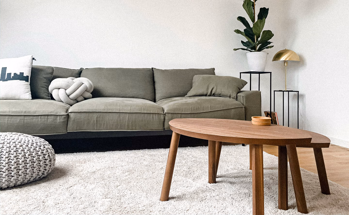 Salon minimaliste avec deux tables basses gigognes en bois de noyer aux plateaux ovales organiques ancrant visuellement l'espace autour d'un canapé kaki profond avec coussin nœud blanc sculptural, tapis blanc moelleux, pouf tricoté gris perle, sellettes noires graphiques avec lampe champignon dorée et figuier lyre illustrant qu'une table basse aux proportions généreuses structure la composition et unifie le mobilier. Salon minimaliste avec deux tables basses gigognes en bois de noyer aux plateaux ovales organiques ancrant visuellement l'espace autour d'un canapé kaki profond avec coussin nœud blanc sculptural, tapis blanc moelleux, pouf tricoté gris perle, sellettes noires graphiques avec lampe champignon dorée et figuier lyre illustrant qu'une table basse aux proportions généreuses structure la composition et unifie le mobilier.