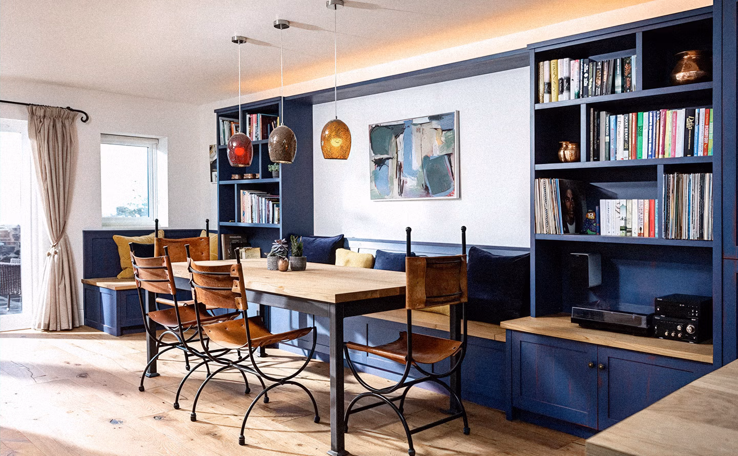 Salle à manger avec bibliothèques verticales intégrées bleu marine encadrant une banquette et table en bois massif avec chaises en fer forgé noir et cuir caramel, trois suspensions en verre ambré rouge et doré attirant le regard vers le haut, parquet bois clair noueux illustrant qu'une stratégie de verticalisation avec mobilier élancé compense les plafonds bas en générant une sensation d'élévation. Salle à manger avec bibliothèques verticales intégrées bleu marine encadrant une banquette et table en bois massif avec chaises en fer forgé noir et cuir caramel, trois suspensions en verre ambré rouge et doré attirant le regard vers le haut, parquet bois clair noueux illustrant qu'une stratégie de verticalisation avec mobilier élancé compense les plafonds bas en générant une sensation d'élévation.