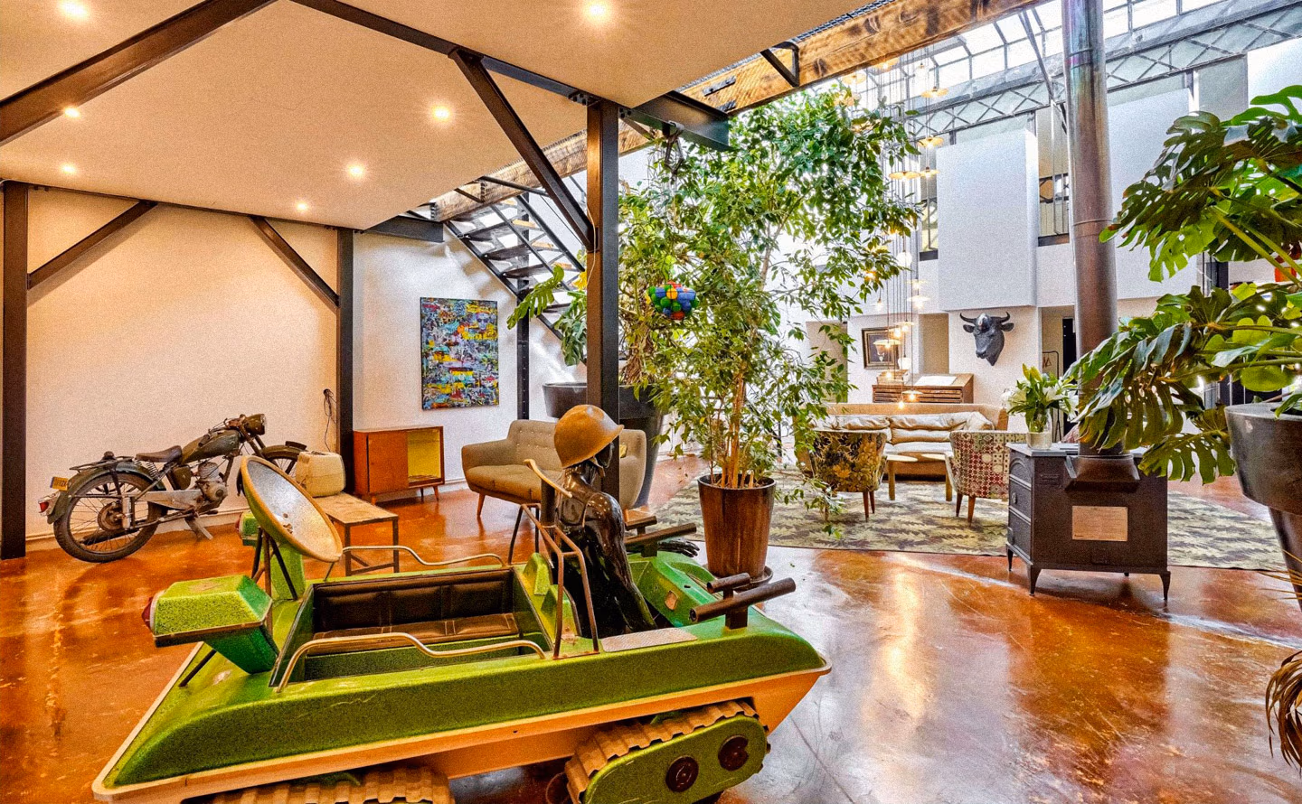 Loft industriel aux poutres apparentes et verrière métallique avec sol béton rouille, accumulation passionnée d'objets chinés incluant moto vintage restaurée, planche de surf ancienne, fauteuils mid-century verts chartreuse et caramel, meuble orange années 60, plantes tropicales luxuriantes en pots terracotta, skateboard, sculptures et tête de taureau illustrant qu'aménager son intérieur doit rester une jubilation créative nourrie par la découverte tangible du monde physique des galeries, antiquaires et marchés aux puces. Loft industriel aux poutres apparentes et verrière métallique avec sol béton rouille, accumulation passionnée d'objets chinés incluant moto vintage restaurée, planche de surf ancienne, fauteuils mid-century verts chartreuse et caramel, meuble orange années 60, plantes tropicales luxuriantes en pots terracotta, skateboard, sculptures et tête de taureau illustrant qu'aménager son intérieur doit rester une jubilation créative nourrie par la découverte tangible du monde physique des galeries, antiquaires et marchés aux puces.