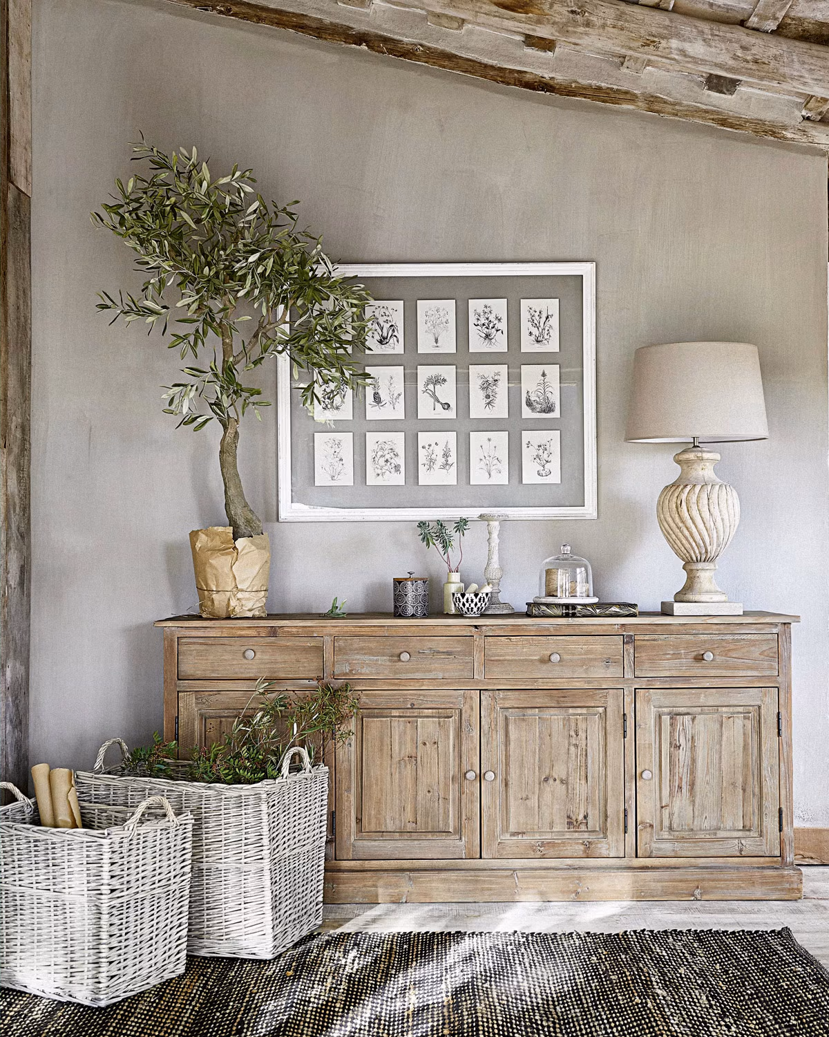 Buffet campagne en bois massif patiné aux tons naturels avec herbier botanique encadré blanc sur fond gris taupe, olivier en pot papier kraft, lampe céramique cannelée crème à abat-jour lin, paniers en osier blanc et poutre apparente ancienne créant une atmosphère campagne apaisante aux tonalités pastel