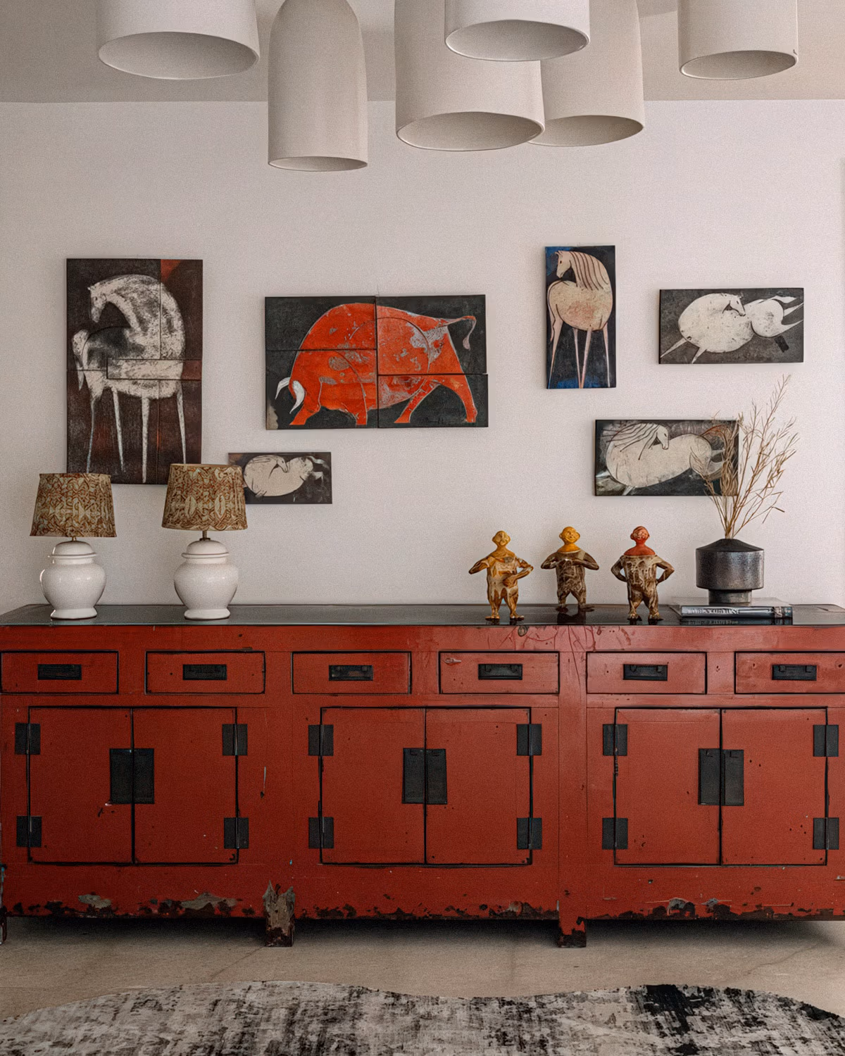 Buffet chinois ancien laqué rouge brique patiné aux ferrures noires avec collection d'œuvres d'art primitif représentant des chevaux stylisés sur murs blanc cassé, lampes en céramique blanche à abat-jour dorés, statuettes tribales en bois et suspensions cylindriques beige créant une composition vintage éclectique sophistiquée
