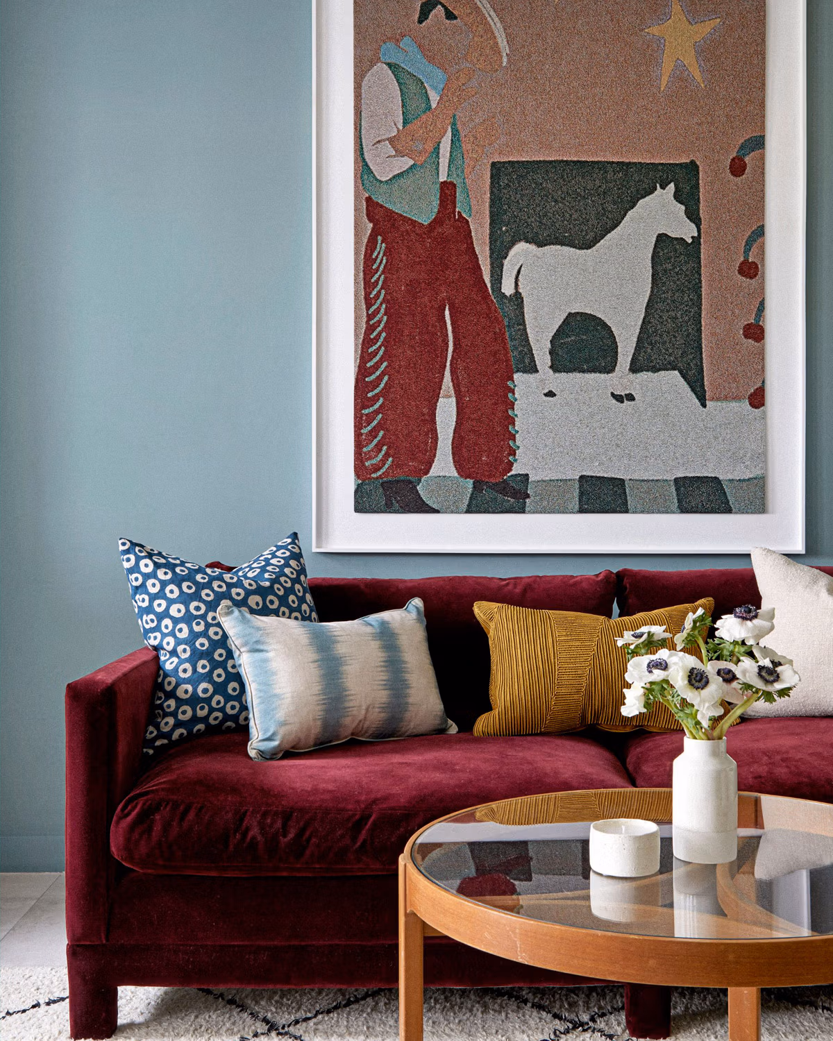 Salon vintage aux murs bleu ciel avec canapé en velours bordeaux profond garni de coussins ethniques bleu marine et moutarde, surmonté d'une œuvre d'art naïf encadrée représentant un cheval blanc et des motifs folkloriques, table basse ronde en bois naturel avec plateau verre et céramiques contemporaines blanches créant une atmosphère seventies sophistiquée