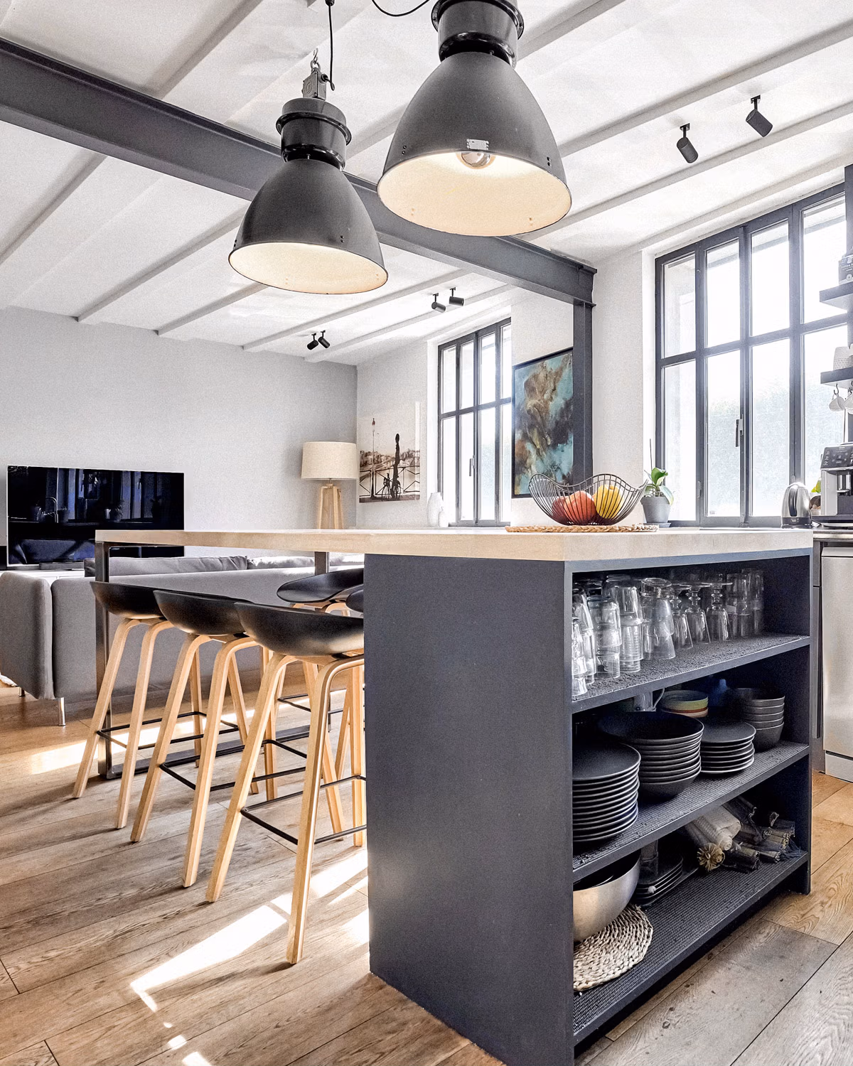 Cuisine ouverte industrielle avec îlot central gris anthracite surmonté d'un plan de travail en pierre claire, suspensions XXL en métal émaillé gris, tabourets de bar scandinaves assise noire et pieds bois naturel, poutrelles métalliques apparentes au plafond blanc, rangements ouverts intégrés à l'îlot et parquet chêne clair baigné de lumière naturelle