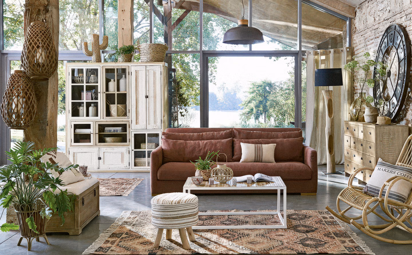 Loft campagne aux larges baies vitrées acier noir donnant sur jardin avec canapé en cuir cognac vieilli, meubles de rangement en bois blanchi patiné, vaisselier vitré ancien, suspension industrielle grise, rocking-chair en rotin naturel, horloge murale surdimensionnée et sol béton ciré créant une atmosphère campagne contemporaine sophistiquée
