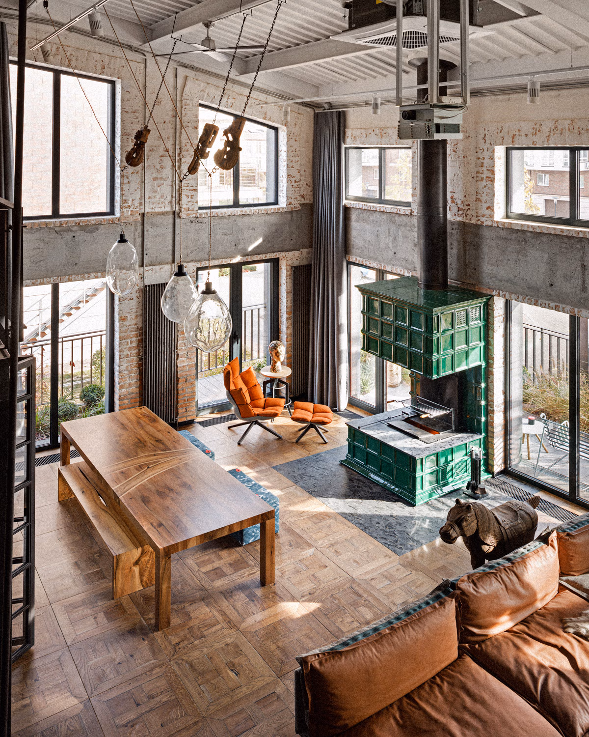Loft industriel à double hauteur avec murs en briques patinées et béton brut, imposant poêle de masse en faïence verte émaillée, fauteuil Husk orange vif, table et banc en bois massif brut, canapé cuir cognac, suspensions en verre soufflé, poutres métalliques apparentes, parquet point de Hongrie et objets vintage suspendus créant des contrastes chromatiques saisissants