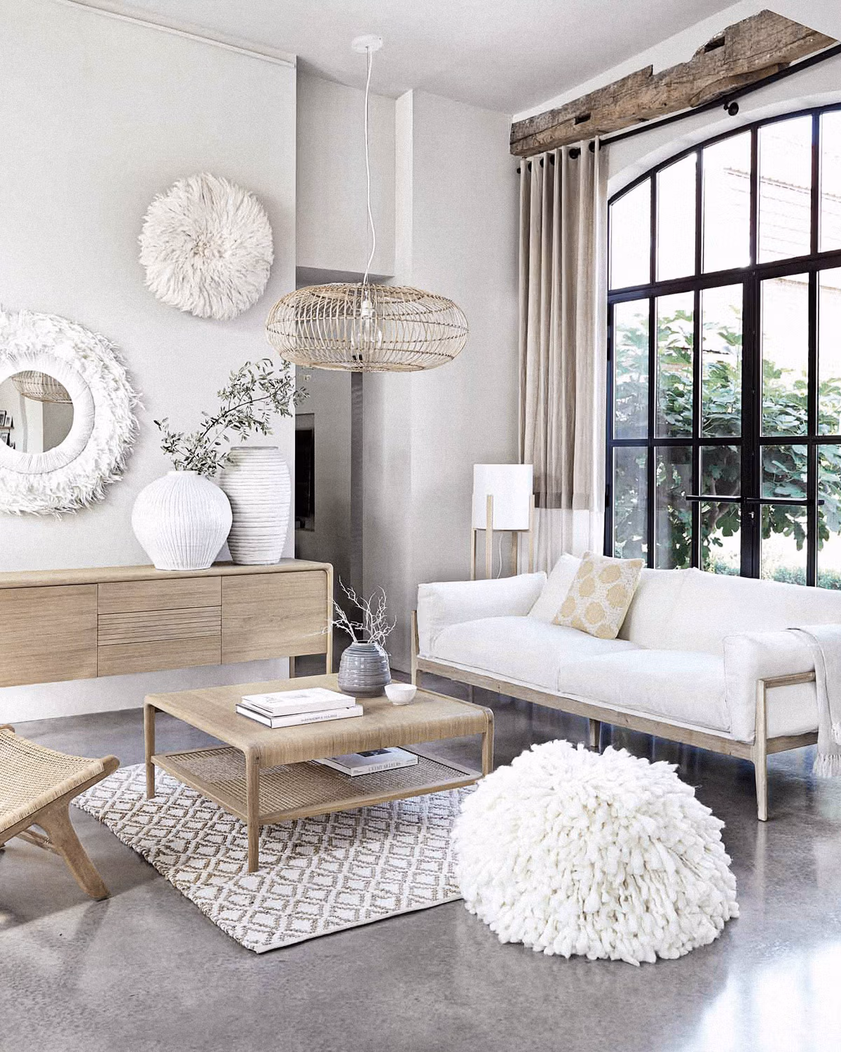 Salon bohème chic aux tons neutres avec canapé en lin blanc sur structure bois, table basse cannée, suspension en rotin naturel, juju hat et miroir en fibres blanches au mur, buffet chêne clair, vases céramiques striées, pouf shaggy laine crème, tapis géométrique beige sur sol béton ciré gris et grande fenêtre cintrée en métal noir laissant entrer la lumière naturelle