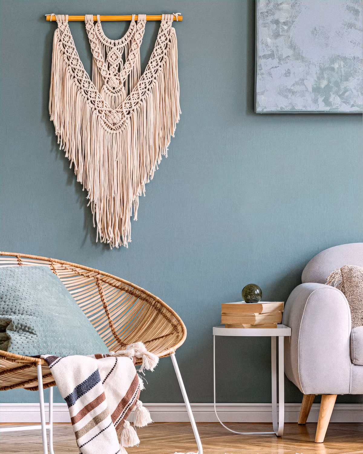 Coin salon bohème avec grande suspension murale en macramé écru aux nœuds complexes et franges sur mur vert sauge, fauteuil Acapulco en rotin naturel garni d'un coussin assorti, plaid rayé à pompons, canapé gris clair aux pieds bois et tableau abstrait dans les tons verts créant une atmosphère artisanale apaisante