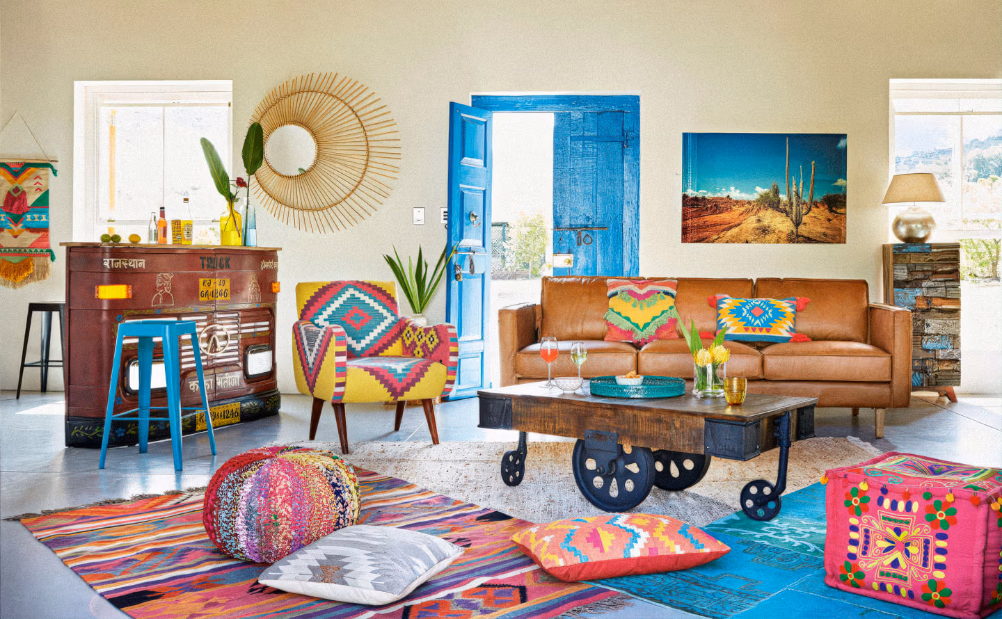 Salon bohème maximaliste avec canapé cuir camel garni de coussins kilim multicolores, fauteuil jaune à motifs aztèques, porte patinée bleu turquoise, meuble bar indien peint style truck art du Rajasthan, table basse industrielle sur roulettes, miroir soleil en bambou, tapis superposés aux géométries vives, poufs brodés et tableau désert créant une explosion chromatique assumée