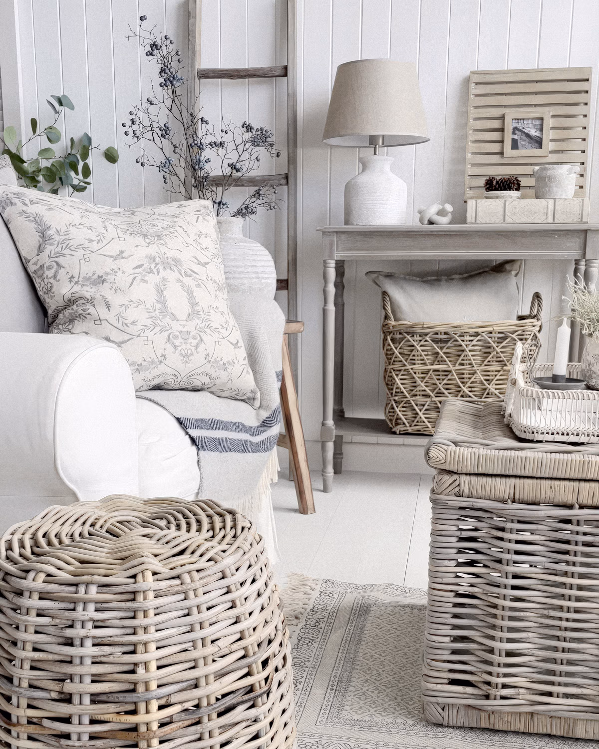 Salon bord de mer épuré avec canapé housse lin blanc, coussins à motifs botaniques gris, paniers en osier grisé empilés, console patinée gris clair, lampe céramique blanche, échelle décorative en bois blanchi, branches de baies séchées bleues et lambris vertical blanc créant une atmosphère côtière tout en retenue
