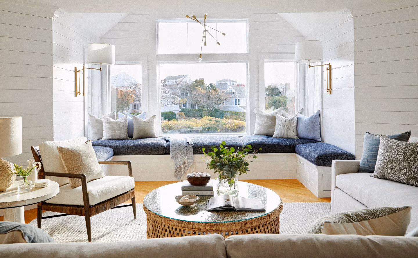 Salon bord de mer style Hamptons avec murs en lambris shiplap blanc, banquette d'angle sous fenêtres garnie de coussins bleu marine et écru, table basse ronde en rotin tressé plateau verre, fauteuils lin naturel, appliques laiton doré et suspension Sputnik, parquet chêne clair et bouquet de feuillage créant une atmosphère de vacances permanente