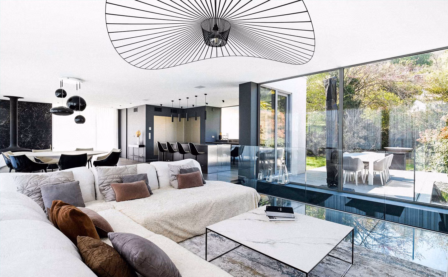 Salon contemporain ouvert avec suspension sculpturale Vertigo de Petite Friture en noir, grand canapé modulable blanc aux assises généreuses garni de coussins velours taupe et terracotta, table basse plateau marbre blanc sur piètement métal noir, immense baie vitrée ouvrant sur jardin arboré et terrasse, cuisine intégrée en arrière-plan