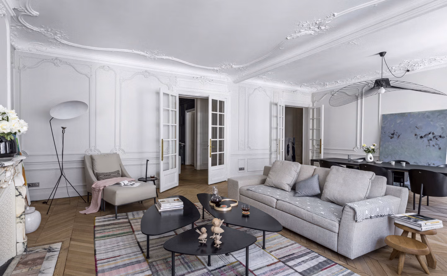 Salon haussmannien aux murs blancs avec moulures sculptées et rosaces de plafond ornementées, parquet en chevrons point de Hongrie, canapé contemporain gris clair lin, fauteuil beige capitonné, tables basses gigognes noires design, ventilateur suspension Vertigo noir, double porte vitrée à carreaux et cheminée marbre blanc créant une atmosphère classique chic épurée moderne