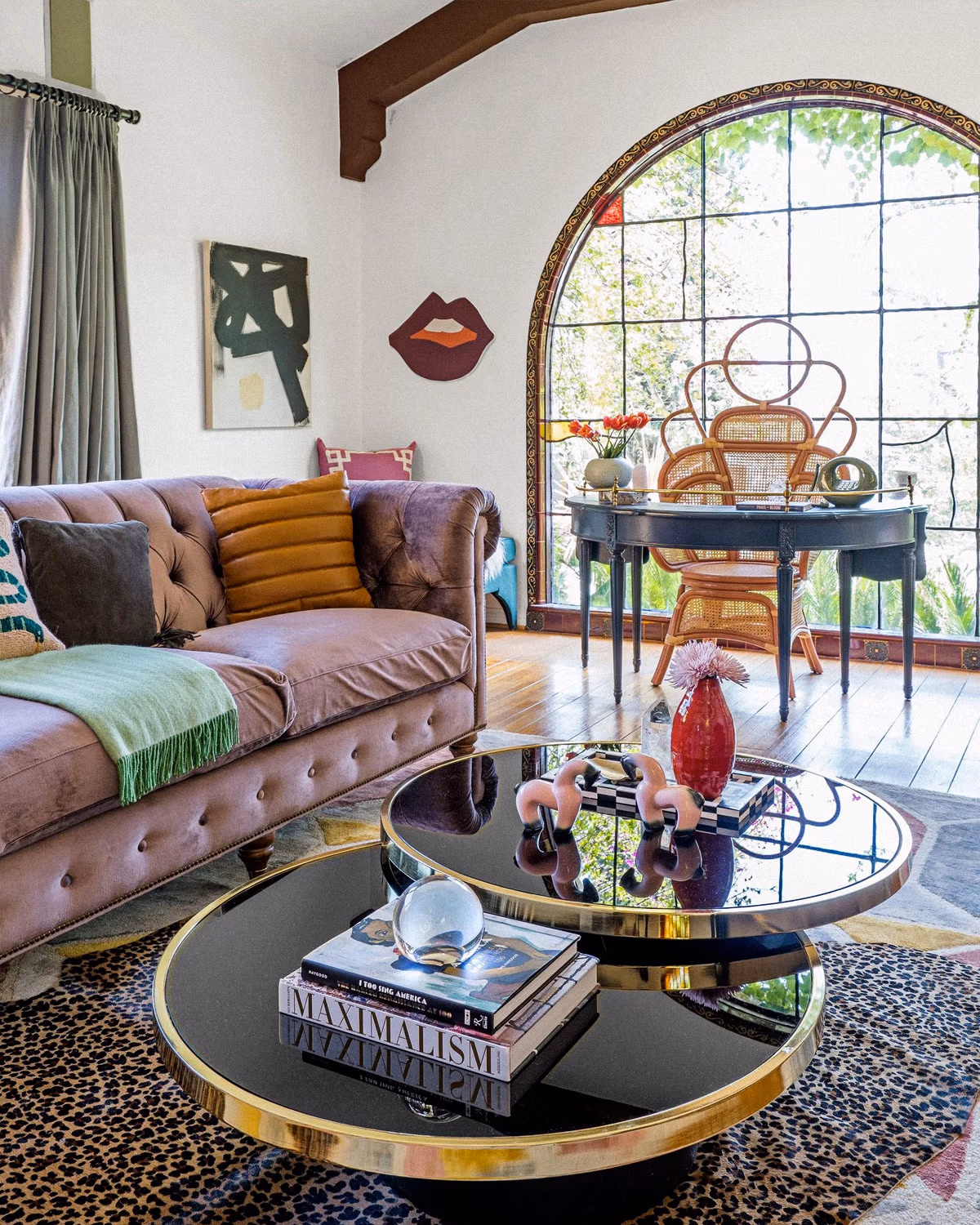 Salon maximaliste éclectique avec canapé Chesterfield capitonné en velours mauve, table basse double plateau verre fumé et laiton doré avec livre Maximalism, chaises Emmanuelle en rotin cannage devant bureau ancien noir, grande fenêtre cintrée hispanique, tapis léopard, sculpture lèvres au mur, coussins dépareillés et objets d'art créant un mélange audacieux d'époques et d'origines