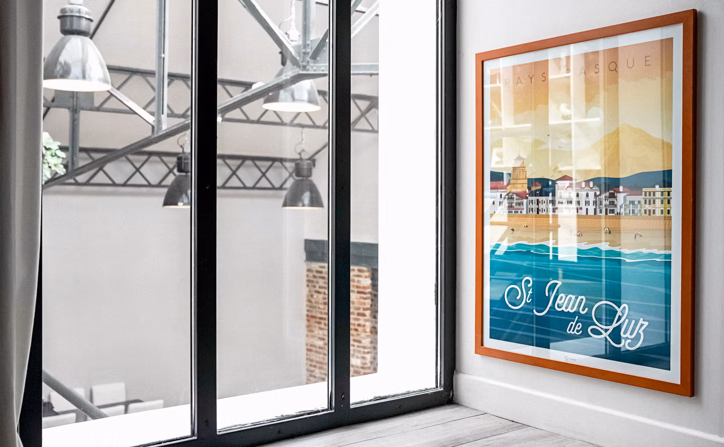 Verrière industrielle à trois panneaux en acier noir donnant sur un espace loft avec charpente métallique apparente et suspensions d'atelier, affiche vintage illustrée Saint-Jean-de-Luz encadrée bois terracotta sur mur blanc, parquet blanchi et briques apparentes en arrière-plan