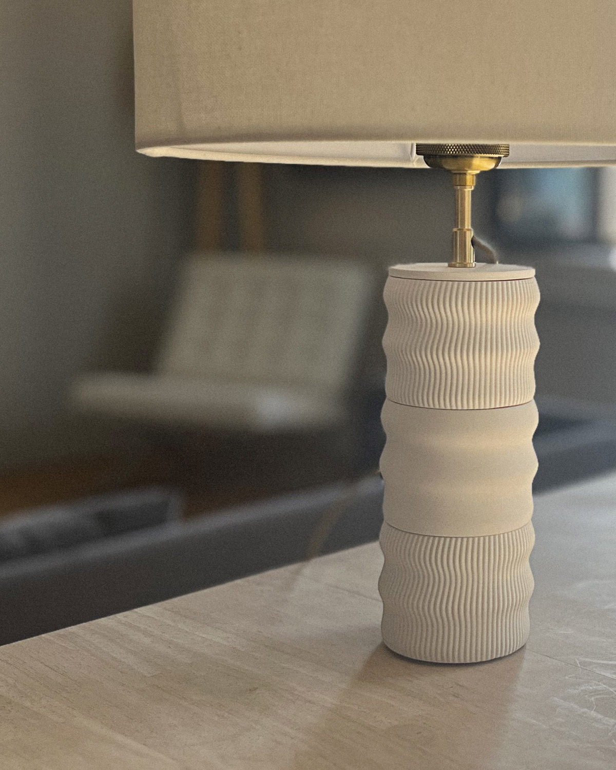 Détail portrait du socle Illari Maison Demaille en configuration Onde triple, stries sinueuses en blanc crème, raccord en laiton doré ciselé au premier plan sous l'abat-jour en lin naturel partiellement visible, posé sur console en chêne clair avec reflet de fauteuil contemporain en arrière-plan Détail portrait du socle Illari Maison Demaille en configuration Onde triple, stries sinueuses en blanc crème, raccord en laiton doré ciselé au premier plan sous l'abat-jour en lin naturel partiellement visible, posé sur console en chêne clair avec reflet de fauteuil contemporain en arrière-plan