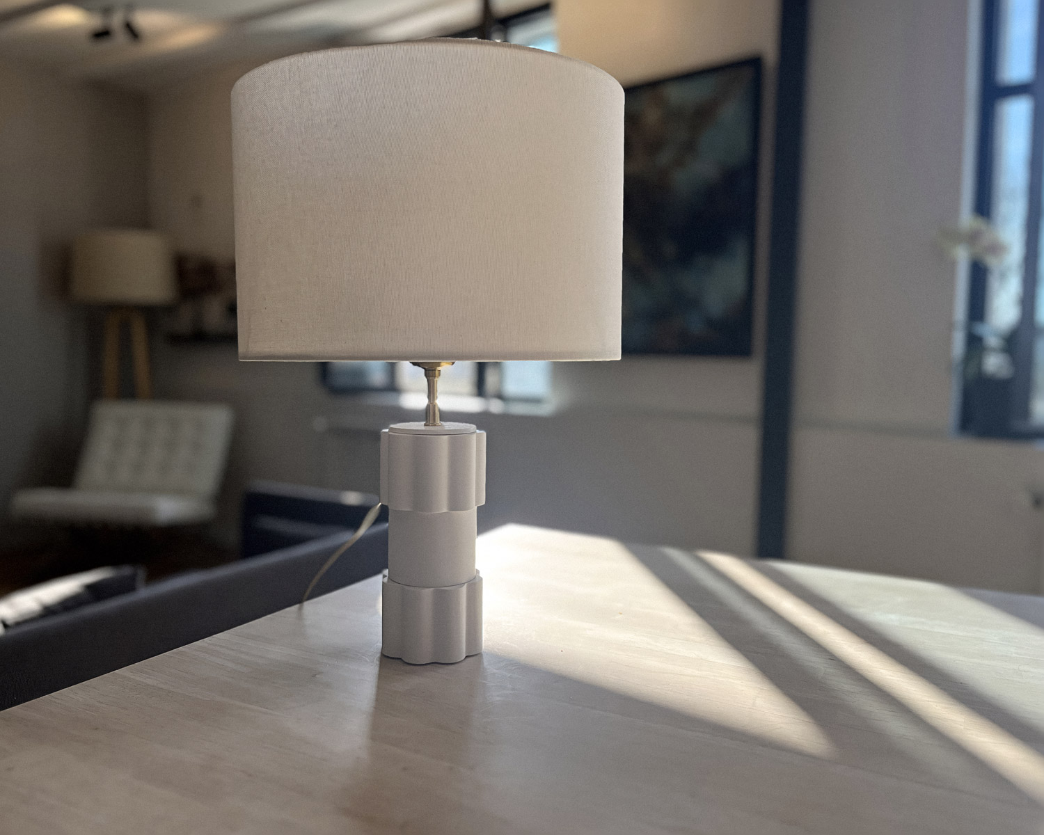 Lampe de table Maison Demaille collection Illari à socle modulable blanc mat et abat-jour en lin crème, posée sur une console en chêne clair baignée de lumière naturelle rasante, dans un intérieur contemporain avec chaise Barcelona et œuvre d'art en arrière-plan Lampe de table Maison Demaille collection Illari à socle modulable blanc mat et abat-jour en lin crème, posée sur une console en chêne clair baignée de lumière naturelle rasante, dans un intérieur contemporain avec chaise Barcelona et œuvre d'art en arrière-plan